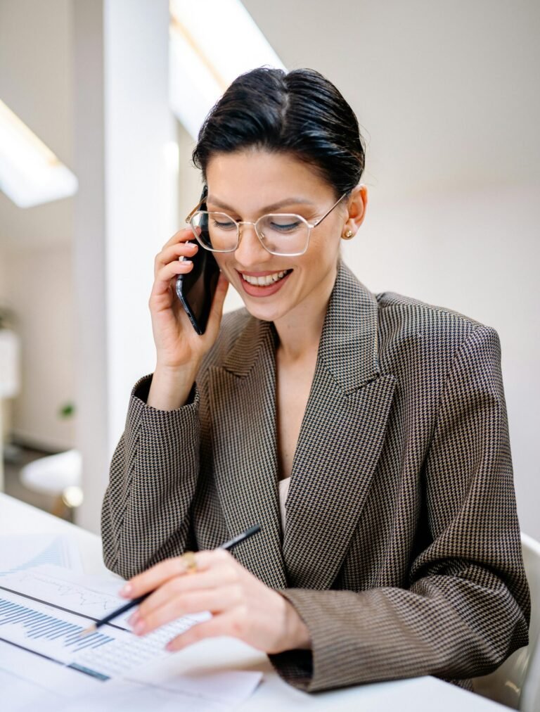 Confident businesswoman wearing glasses, talking on phone while reviewing documents in modern office setting.